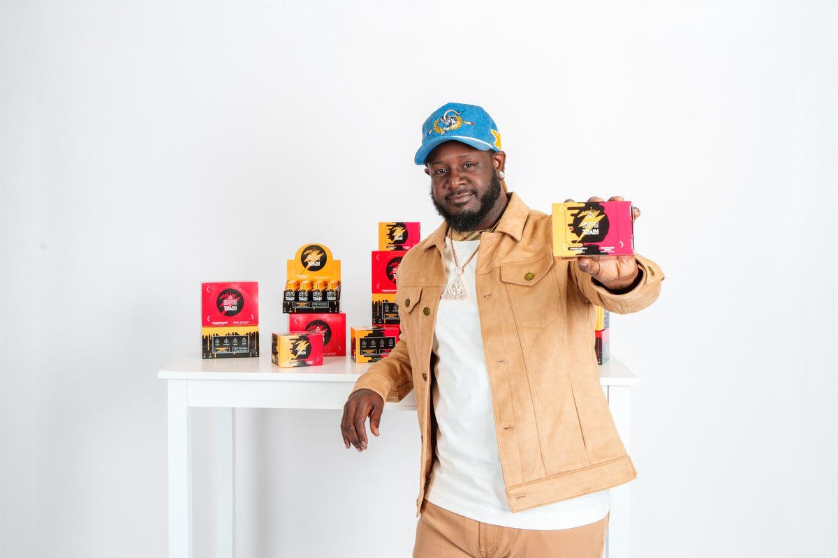 T-Pain holding a Good Game by T-Pain energy shot box beside a table of Strawberry Surge and Orange Vanilla Vortex 12-packs